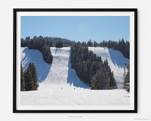 This fine art photography print shows winter in Vail, Colorado at Vail Ski Resort. From the Wildwood Express ski lift, you’ll see stunning views of skiers at the bottom of the Look Ma and Challenge black diamond ski runs. The scene is framed by blue sky and pine trees amongst the Rocky Mountains.