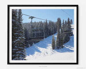 This fine art photography print shows winter in Vail, Colorado at Vail Ski Resort. From the Wildwood Express ski lift is a striking view of an empty ski chair lift car set against the snow-covered Rocky Mountains. Clear blue skies are pictured with the sun shining down on pine trees that cast a shadow on the slopes.