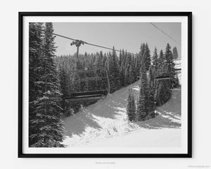 This black and white fine art photography print shows winter in Vail, Colorado at Vail Ski Resort. From the Wildwood Express ski lift is a striking view of an empty ski chair lift car set against the snow-covered Rocky Mountains. Clear blue skies are pictured with the sun shining down on pine trees that cast a shadow on the slopes.