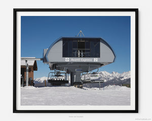 This fine art photography print shows winter in Vail, Colorado at Vail Ski Resort. At the top of the mountain, the Avanti Express ski lift is flanked by a clear blue sky and the snow-capped Rocky Mountains.