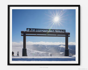 This fine art photography print shows winter in Vail, Colorado at Vail Ski Resort. Two skiers stand next to the Legendary Back Bowls sign. The sun is shining, and the skies are blue with fluffy clouds lining the horizon and the Colorado Rockies in the distance.