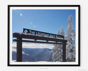This fine art photography print shows winter in Vail, Colorado at Vail Ski Resort. The Legendary Back Bowls sign stands tall with snow-covered pine trees next to the landmark. The sun is shining, and the skies are blue with fluffy clouds lining the horizon and the Colorado Rockies in the distance.
