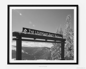 This black and white fine art photography print shows winter in Vail, Colorado at Vail Ski Resort. The Legendary Back Bowls sign stands tall with snow-covered pine trees next to the landmark. The sun is shining, and the skies are blue with fluffy clouds lining the horizon and the Colorado Rockies in the distance.