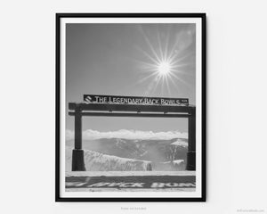 This black and white fine art photography print shows winter in Vail, Colorado at Vail Ski Resort. The Legendary Back Bowls sign stands tall with the shadows of its name cast down in the snow.  With the sun shining overhead, fluffy white clouds line the horizon.