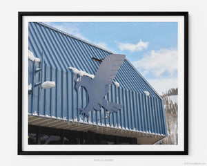 This fine art photography print shows winter in Vail, Colorado at Vail Ski Resort. The sign of the blue eagle on the side of the Eagle Bahn Gondola in Lionshead Village is shown. Small icicles are shown suspended from the gondola and a glimpse of the side of a snow-covered Vail Mountain is shown in the distance.