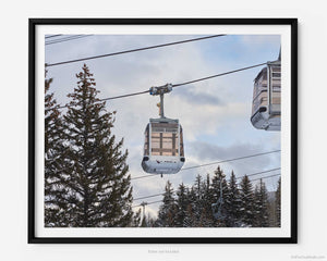 This fine art photography print shows winter in Vail, Colorado at Vail Ski Resort. An empty cable car, suspended from the the Eagle Bahn Gondola, ascends the mountain from Lionshead Village with snow covered pine trees and a partially cloudy sky  in the distance.