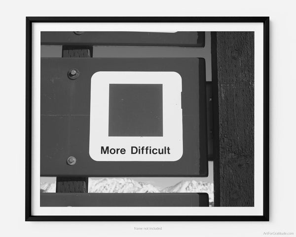 This black and white fine art photography print shows winter in Vail, Colorado at Vail Ski Resort. The air is crisp and cool with a blue (intermediate) ski slope sign that reads "more difficult", near the top of Wildwood Express ski lift.