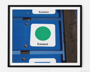 This fine art photography print shows winter in Vail, Colorado at Vail Ski Resort. The air is crisp and the sky is blue while you take in a green (beginner) ski slope sign that reads “easiest”, near the top of Wildwood Express ski lift.