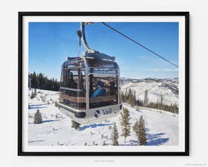 This fine art photography print shows winter in Vail, Colorado at Vail Ski Resort. On a sunny day, the skies are blue with a few scattered clouds resting across the horizon as a passing cable car filled with eager skiers heads up the side of a mountain for a day of fun in the snow-covered Rocky Mountains. 