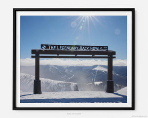 This fine art photography print shows winter in Vail, Colorado at Vail Ski Resort. The Legendary Back Bowls sign stands tall. The sun is shining, and the skies are blue with fluffy clouds lining the horizon and the Colorado Rockies in the distance.