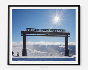 This fine art photography print shows winter in Vail, Colorado at Vail Ski Resort. Two skiers stand next to the Legendary Back Bowls sign. The sun is shining, and the skies are blue with fluffy clouds lining the horizon and the Colorado Rockies in the distance. 