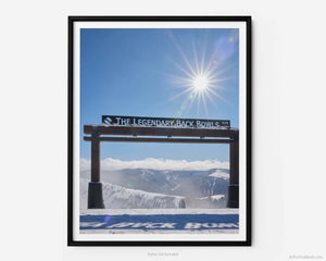 This fine art photography print shows winter in Vail, Colorado at Vail Ski Resort. The Legendary Back Bowls sign stands tall. The sun is shining, and the skies are blue with fluffy clouds lining the horizon and the Colorado Rockies in the distance.