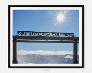 This fine art photography print shows winter in Vail, Colorado at Vail Ski Resort. The Legendary Back Bowls sign stands tall. The sun is shining, and the skies are blue with fluffy clouds lining the horizon and the Colorado Rockies in the distance.