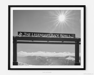 This black and white fine art photography print shows winter in Vail, Colorado at Vail Ski Resort. The Legendary Back Bowls sign stands tall. The sun is shining, and the skies are blue with fluffy clouds lining the horizon and the Colorado Rockies in the distance.