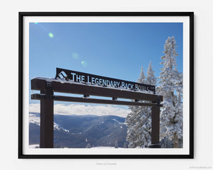  This fine art photography print shows winter in Vail, Colorado at Vail Ski Resort. The Legendary Back Bowls sign stands tall with snow-covered pine trees next to the landmark. The sun is shining, and the skies are blue with fluffy clouds lining the horizon and the Colorado Rockies in the distance.