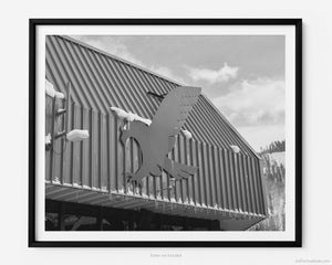 This black and white fine art photography print shows winter in Vail, Colorado at Vail Ski Resort. The sign of the blue eagle on the side of the Eagle Bahn Gondola in Lionshead Village is featured with small icicles suspended from the gondola and a glimpse of a snow-covered Vail Mountain in the distance.