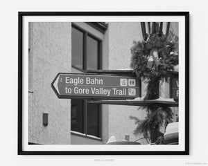 This black and white fine art photography print shows winter in Vail, Colorado at Vail Ski Resort. A sign in Lionshead Village points to the iconic Eagle Bahn Gondola and the Gore Valley Trail.  