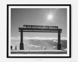 This black and white fine art photography print shows winter in Vail, Colorado at Vail Ski Resort. Two skiers stand next to the Legendary Back Bowls sign planning their next run. The sun is shining, and the skies are blue with fluffy clouds lining the horizon and the Colorado Rockies in the distance. 