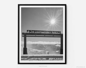 This black and white fine art photography print shows winter in Vail, Colorado at Vail Ski Resort. The Legendary Back Bowls sign stands tall with the shadows of its name cast down in the snow.  With the sun shining overhead, fluffy white clouds line the horizon. 