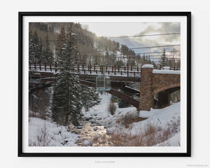 This fine art photography print shows winter in Vail, Colorado at Vail Ski Resort. The iconic Eagle Bahn Gondola moves over Gore Creek at sunset with the snow-covered mountain in the distance. The bridge leading into Lionshead is pictured with sun beams reflecting off of the water in the creek. 