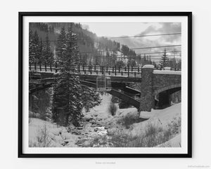 This black and white fine art photography print shows winter in Vail, Colorado at Vail Ski Resort. The iconic Eagle Bahn Gondola moves over Gore Creek at sunset with the snow-covered mountain in the distance. The bridge leading into Lionshead is pictured with sun beams reflecting off of the water in the creek. 