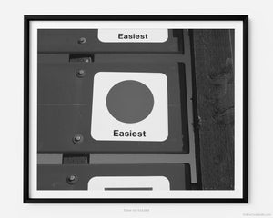 This black and white fine art photography print transports you to winter at Vail Ski Resort in Vail, Colorado. The air is crisp and the sky is blue while you take in a green (beginner) ski slope sign that reads “easiest”, near the top of Wildwood Express ski lift.