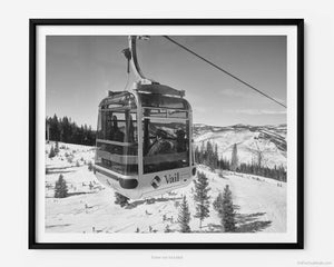 This black and white fine art photography print shows winter in Vail, Colorado at Vail Ski Resort. On a sunny day, the skies are blue with a few scattered clouds resting across the horizon as a passing cable car filled with eager skiers heads up the side of a mountain for a day of fun in the snow-covered Rocky Mountains.