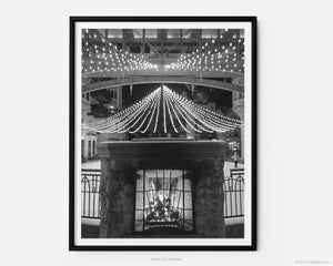 This black and white fine art photography print shows winter in Vail, Colorado at Vail Ski Resort. Located in Vail Square, Lionshead Village, the photo showcases a cozy winter scene of a blazing fireplace set against an ice-skating rink adorned with enchanting Christmas lights.